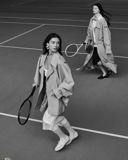 Two models in trench coats carrying rackets on a tennis court, in black and white.