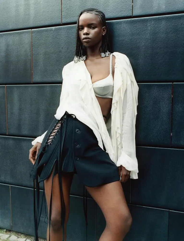 A model in a white shirt and laced black skirt against dark panelling.