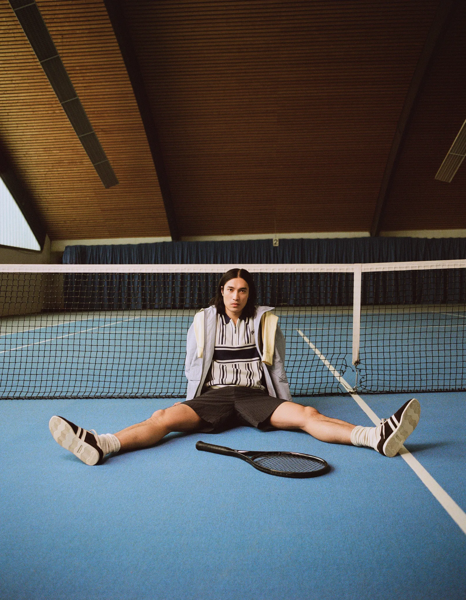 A model in a striped knit and shorts seated on a blue indoor tennis court beside a racket, shot for Schön Magazine.